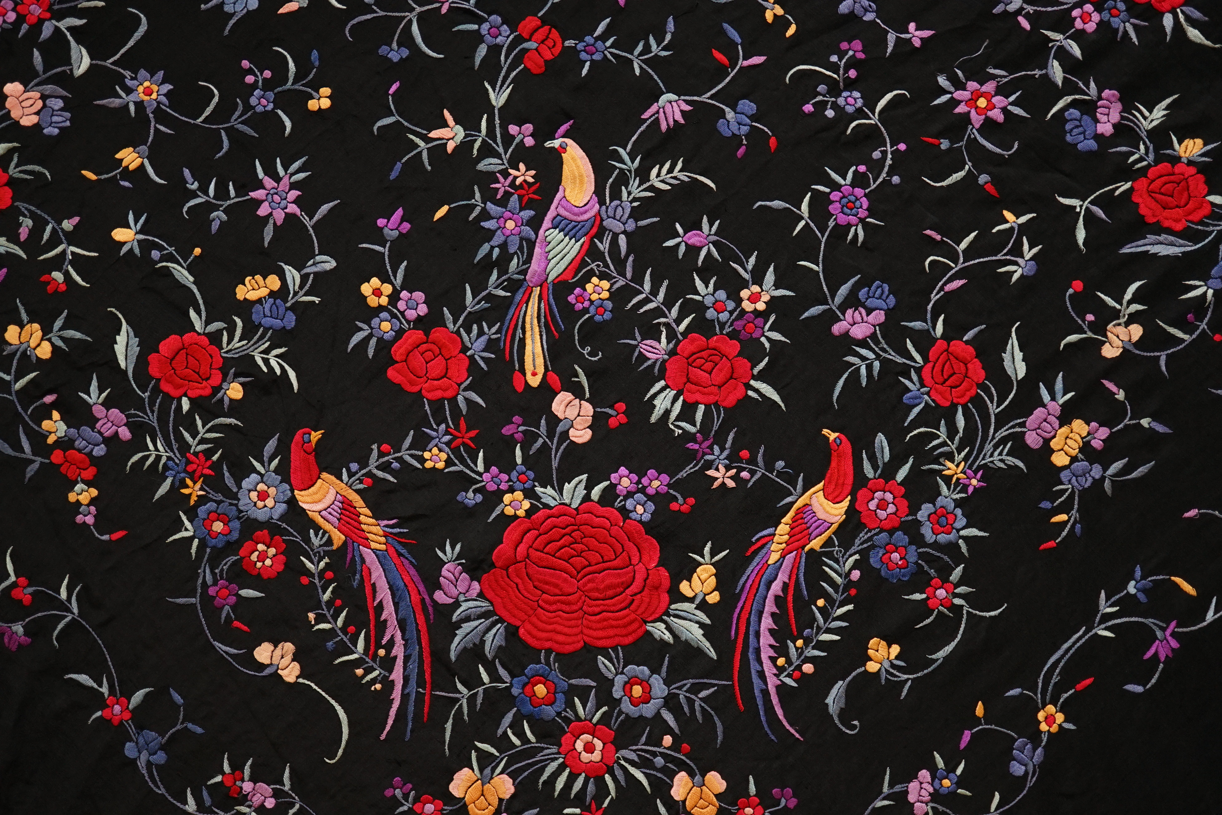 An early 20th century Chinese black silk fringed shawl with polychrome silk embroidery, embroidered with meandering floral design, the corners embroidered with three brightly coloured birds of paradise, large red roses a